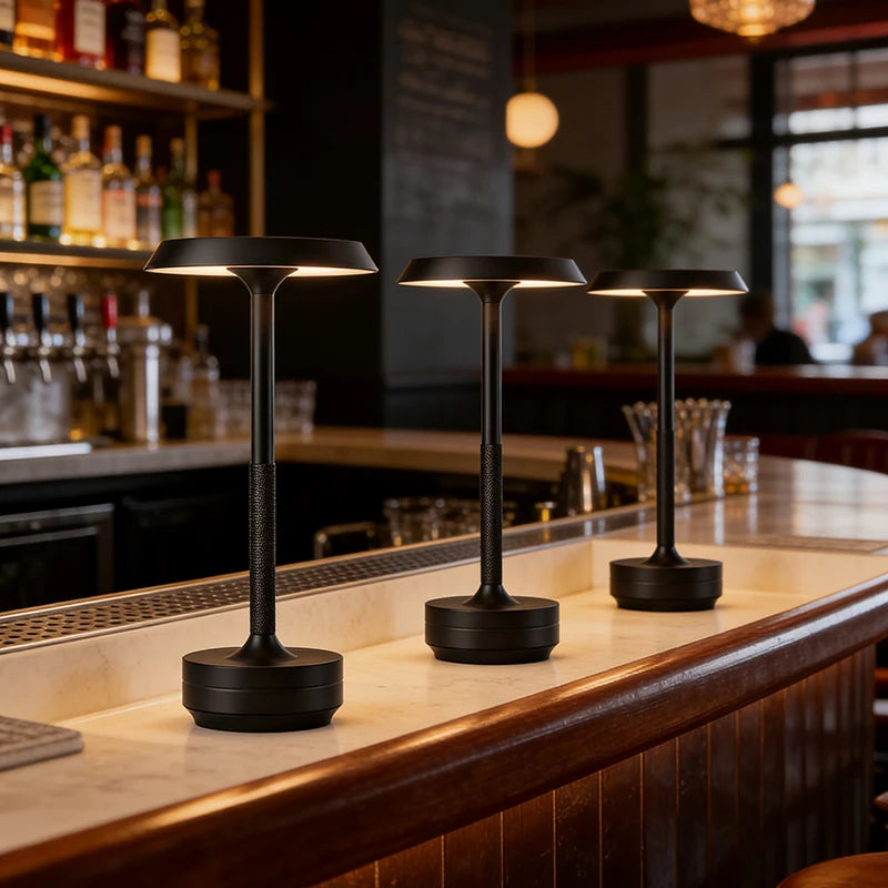  Three black rechargeable table lamps lined up on a marble bar counter in an upscale cocktail bar, glowing with warm ambient light — "Disc & Stem" by CL House