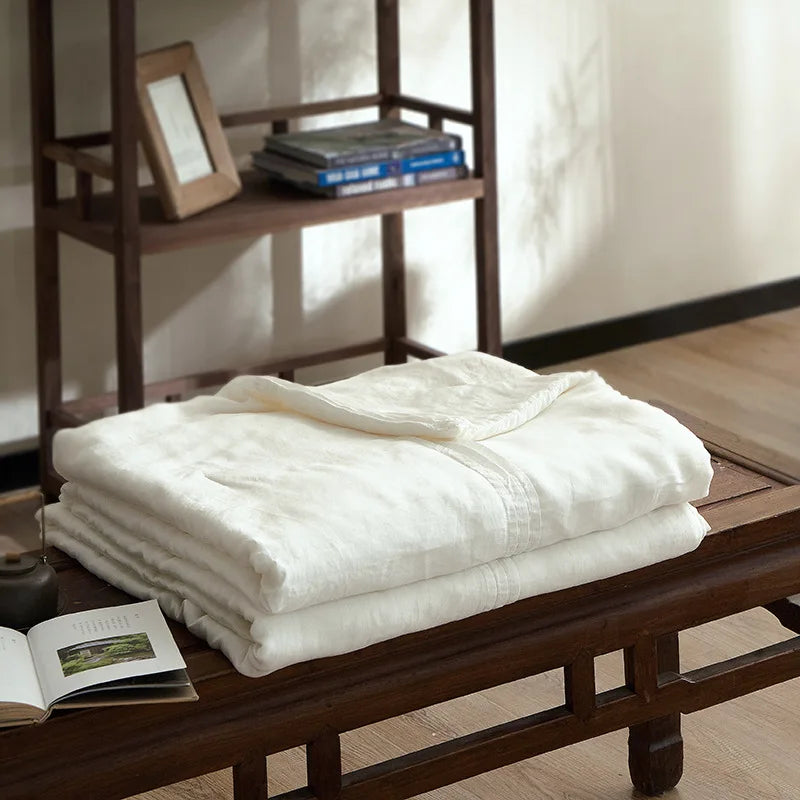  Ivory white quilted linen cotton summer throw blanket, neatly folded on a dark wood bench in a japandi-style interior with wooden shelving and open book