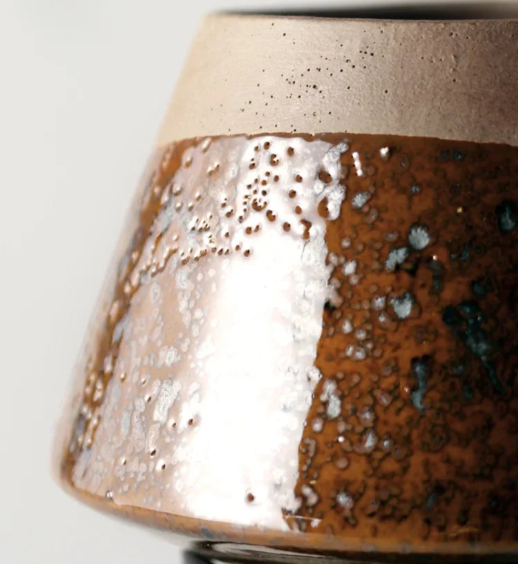Close-up detail of amber reactive glaze ceramic plant pot showing rich brown, silver and teal glaze texture with speckled sand beige rim, white background