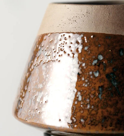 Close-up detail of amber reactive glaze ceramic plant pot showing rich brown, silver and teal glaze texture with speckled sand beige rim, white background