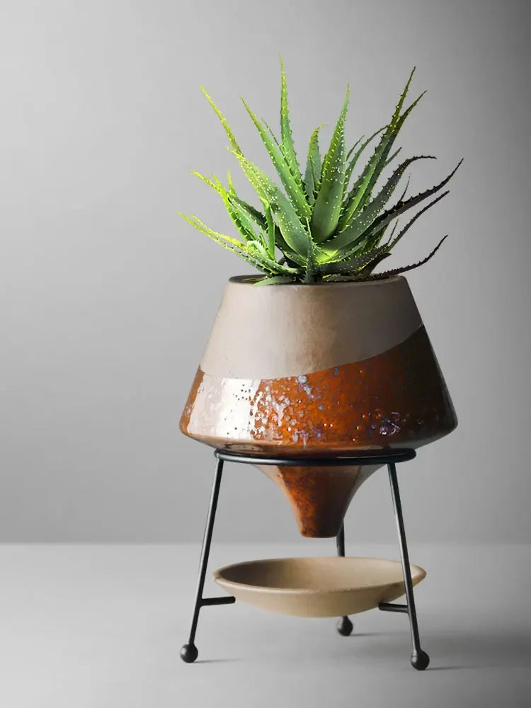  Amber glaze ceramic cone plant pot on black iron tripod stand holding a spiky aloe vera plant, rich brown reactive glaze with sand beige top, grey background