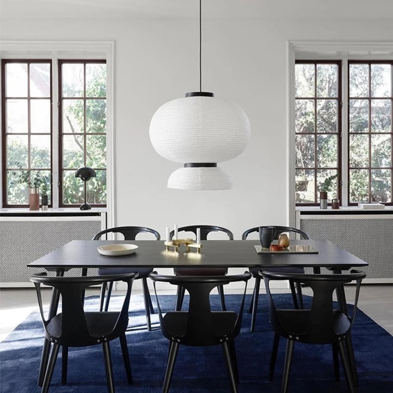 : Rice & Ripple paper pendant light hanging above a large black dining table with black chairs in a bright white dining room with tall windows and a navy blue rug