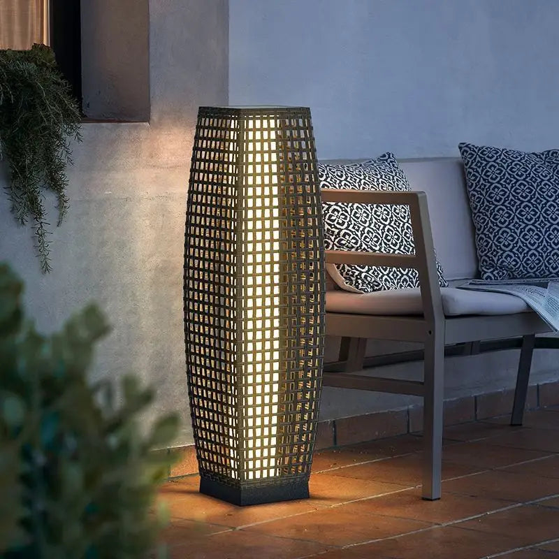 Tall dark wicker solar floor lantern glowing with warm amber light on a covered patio beside a grey outdoor sofa with blue patterned cushions at dusk