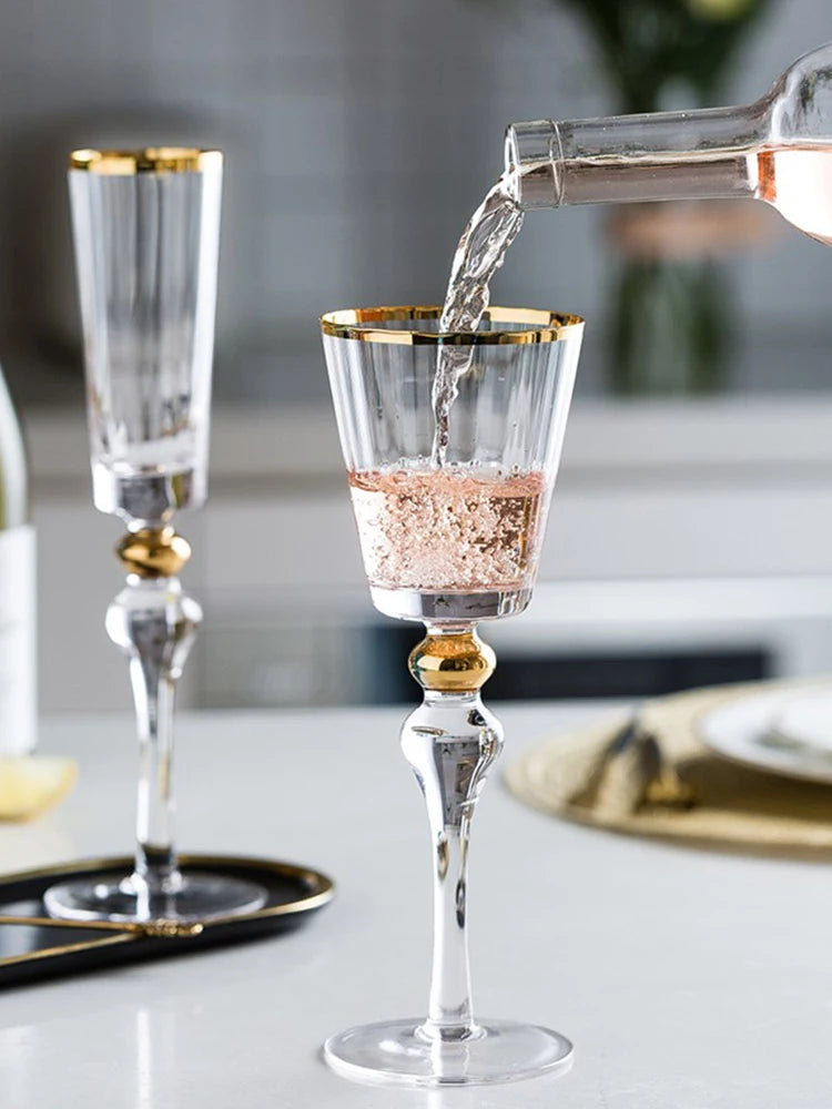 Hand holding a "Golden Hour" crystal wine glass filled with red wine alongside a champagne flute, gold filigree rim detail — CL House Drinkware collection