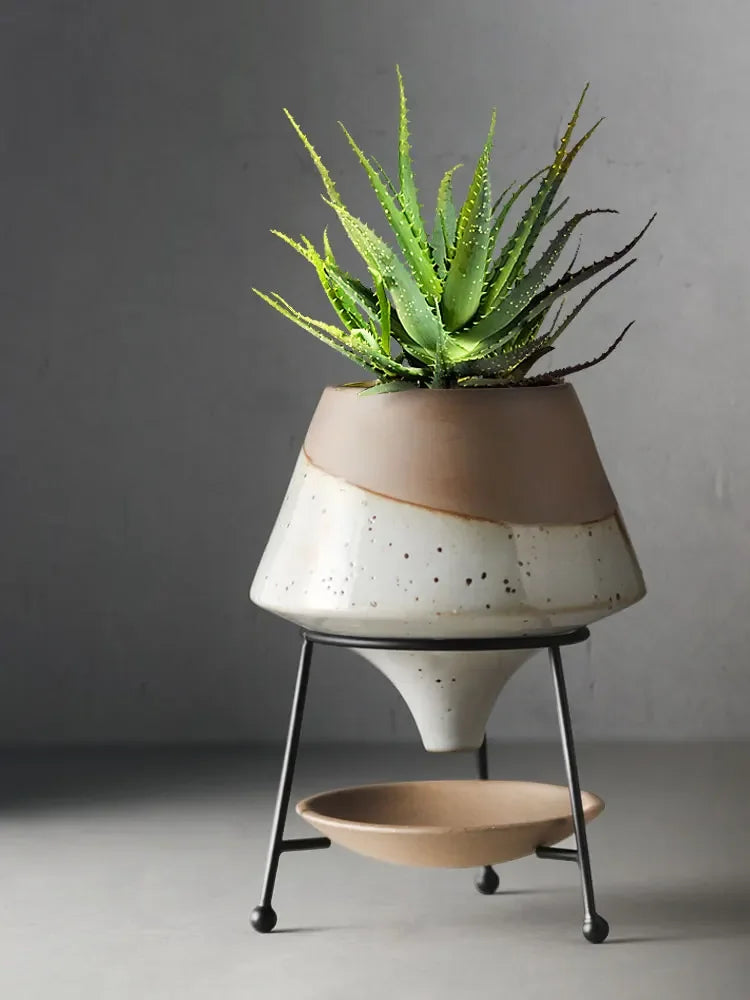 Speckled white reactive glaze ceramic cone plant pot on black iron tripod stand holding an aloe vera plant, speckled white and terracotta two-tone finish, grey background