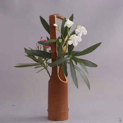 Handmade dark bamboo sculptural vase with rattan handle, styled with white oleander blooms, pink flowers and long green leaves against a light grey background