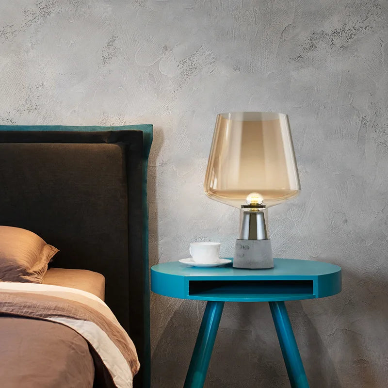 "Smoke & Cement" glass table lamp with amber shade styled on a teal side table next to a dark upholstered bed against a concrete wall