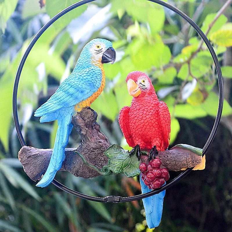 "Perch Duo" resin parrot hanging ring ornament in Scarlet & Azure — close-up detail of blue and red macaws on driftwood branch with leaf and berry cluster, garden foliage background