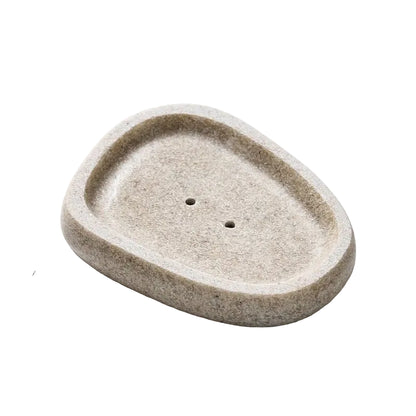 Oval pebble-shaped stone-texture resin soap dish in warm sand beige with two drainage holes, top angle view, white background