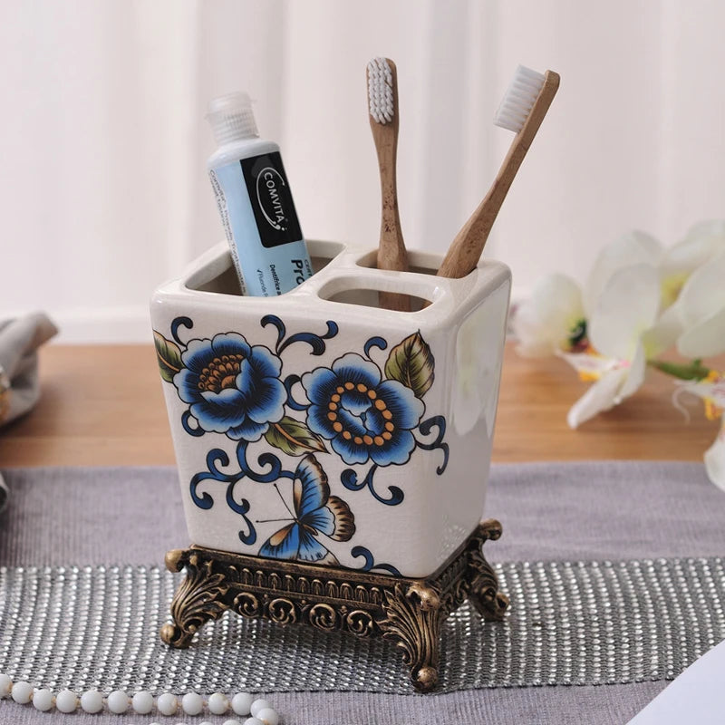  White ceramic toothbrush holder with blue chinoiserie rose and butterfly pattern, ornate bronze metal base, two bamboo toothbrushes and toothpaste displayed, lifestyle background