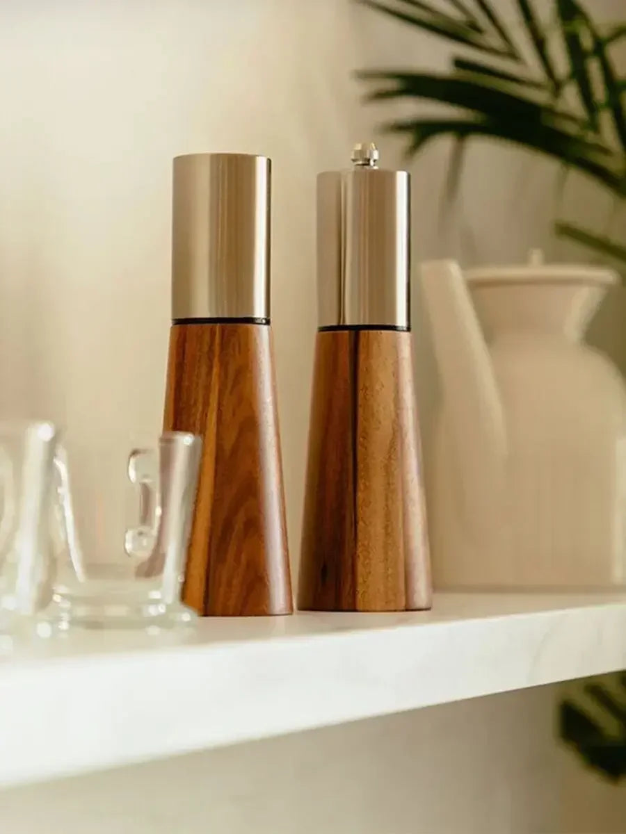 Acacia Twist salt and pepper mill duo styled on a white shelf with glass vessels and a ceramic vase, with a tropical palm leaf in the background