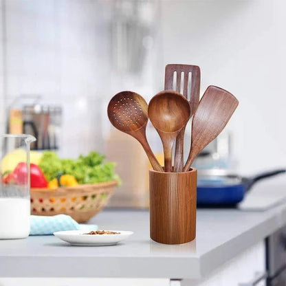  Teak wood cooking utensil set displayed in matching cylindrical teak holder on a white kitchen countertop — skimmer, ladle, slotted spatula and wok spatula fanned out, fresh vegetables and fruit basket in soft-focus background — lifestyle hero shot of the Teak Grove Utensil Set by CL House