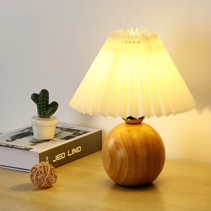 "Pleat & Wood" pleated table lamp with off white shade and round wood sphere base glowing warmly, styled on a wooden surface with a mini cactus and rattan ball