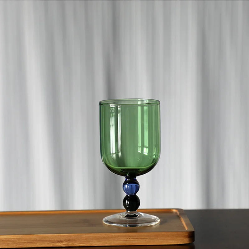 "Chromatic Orb" glass goblet in forest green with two-tone blue and black bead stem, on a wooden tray against a white curtain background