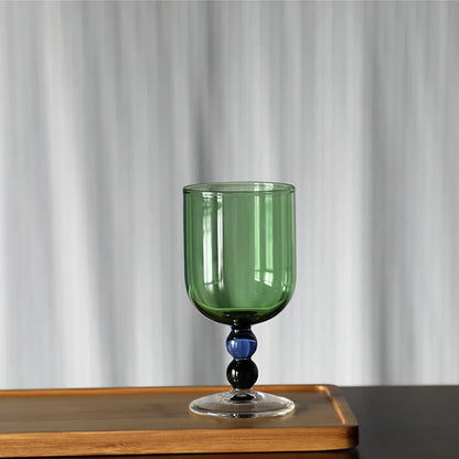 "Chromatic Orb" glass goblet in forest green with two-tone blue and black bead stem, on a wooden tray against a white curtain background
