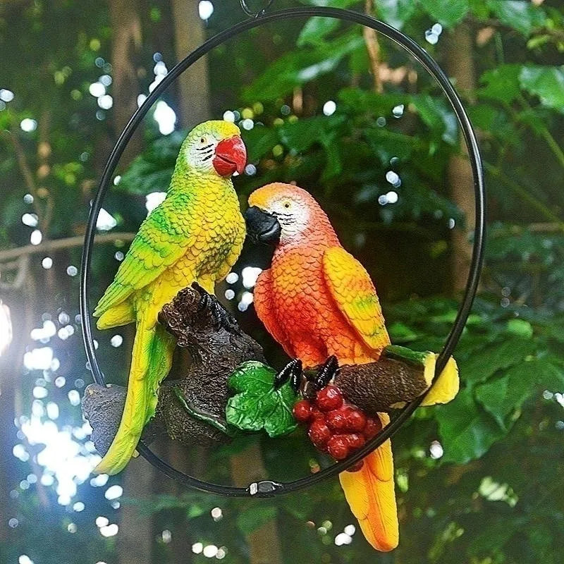 "Perch Duo" resin parrot hanging ring ornament in Emerald & Amber — night garden setting with bokeh lights, green and orange parrots facing each other on driftwood branch