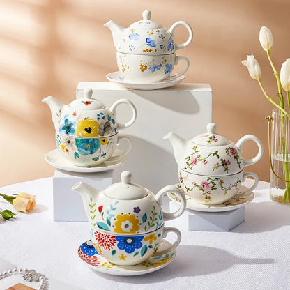 Four floral porcelain tea for one sets displayed on white geometric blocks with flowers and tulips, showing all four designs: blue chicory, watercolour blooms, pink roses and folk flowers, warm beige background