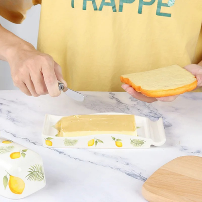 Hands spreading butter on a slice of bread using a knife, with a lemon-patterned ceramic butter dish tray on a marble surface and the domed lid set aside.