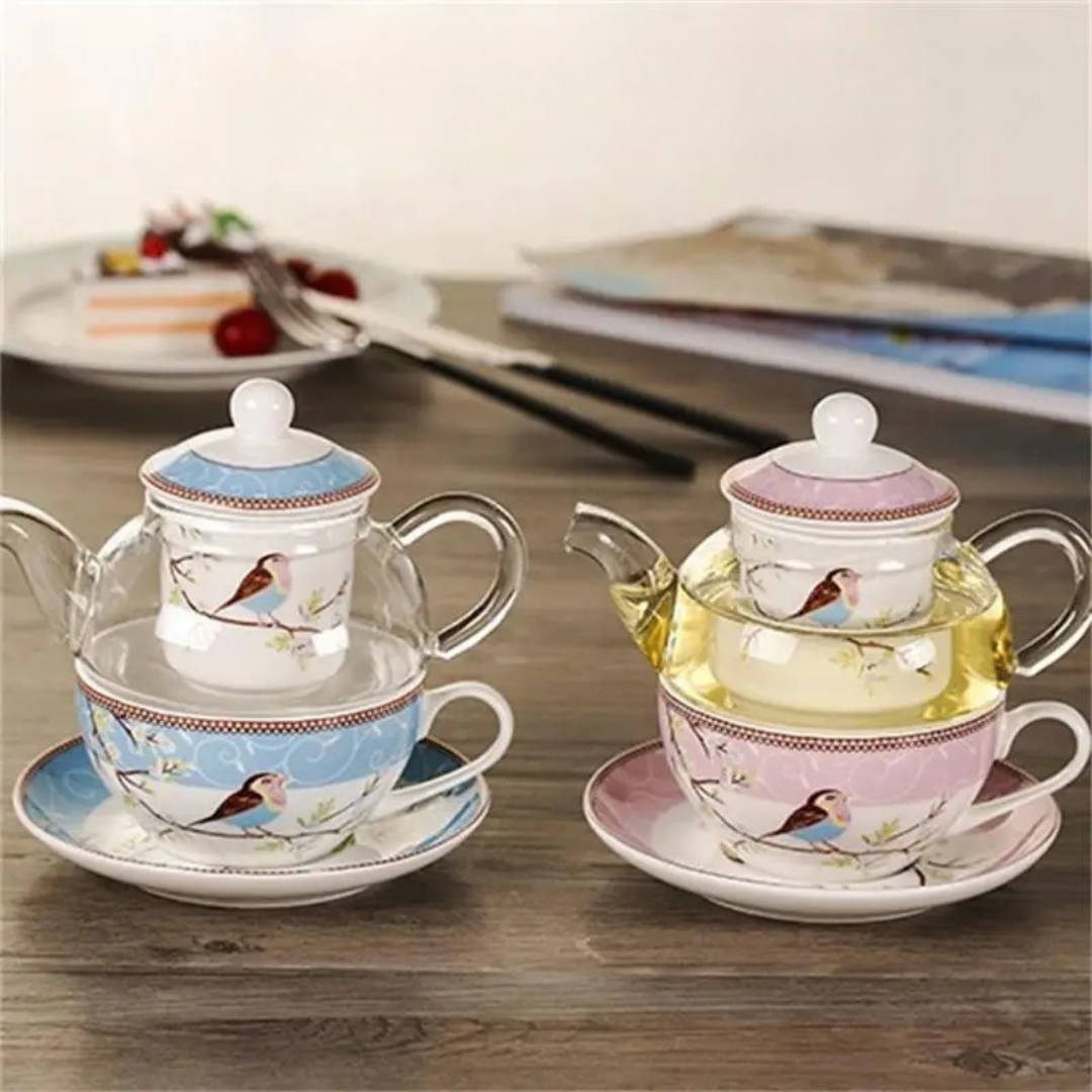 Blue and pink "Happy Bird" tea for one sets side by side on a wooden table — each with a glass teapot, porcelain infuser cup and saucer, bird and blossom pattern, with cake plate and magazine in the background.