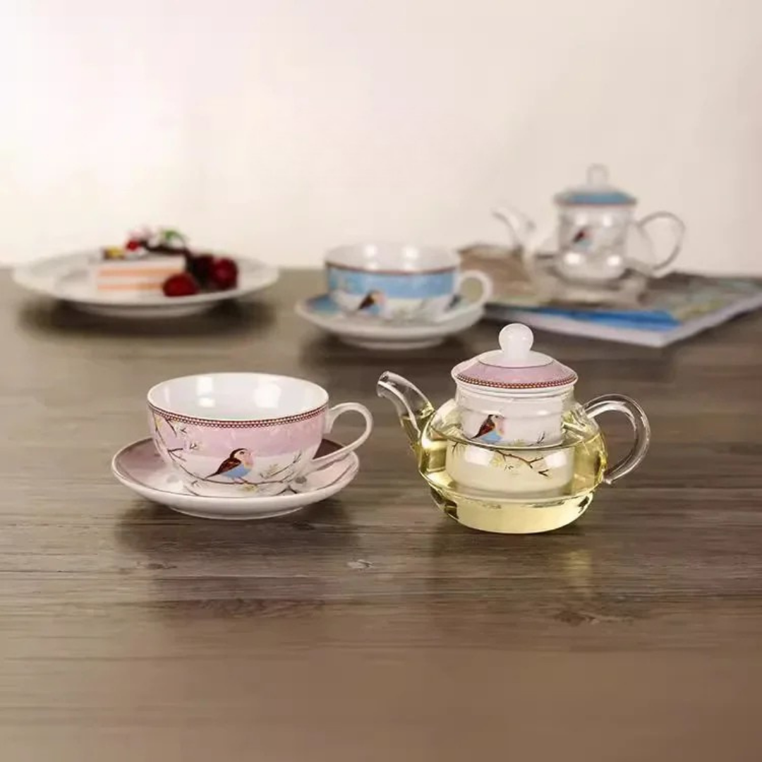 Pink and blue "Happy Bird" tea for one sets styled on a wooden table — pink porcelain cup with saucer and glass teapot filled with golden tea in the foreground, blue set and cake plate in the background.