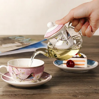 Hand pouring golden herbal tea from a pink "Happy Bird" glass teapot into a porcelain cup and saucer on a wooden table, with a slice of cake and magazine softly blurred in the background.