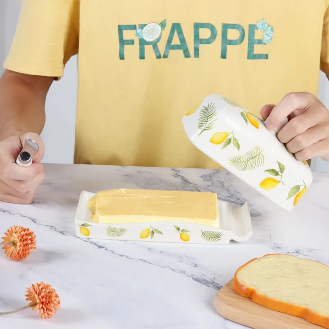 Hands lifting the domed lid of a lemon-patterned ceramic butter dish revealing a block of butter on a marble surface, with dried orange flowers and a slice of bread alongside.