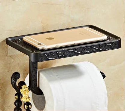  Wall-mounted matte black toilet paper holder with engraved arabesque pattern, gold accent details and phone shelf, smartphone resting on top, marble wall background