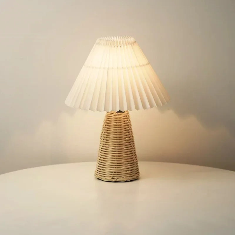Rattan & Pleat table lamp with woven rattan cone base and wide layered pleated shade in off-white, lit with warm light on a white table against a soft grey wall