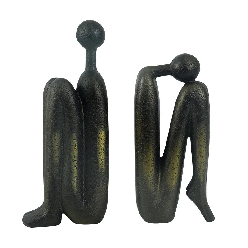 Pair of dark resin abstract human sculptures standing side by side against a white background, with one figure upright and the other leaning forward.