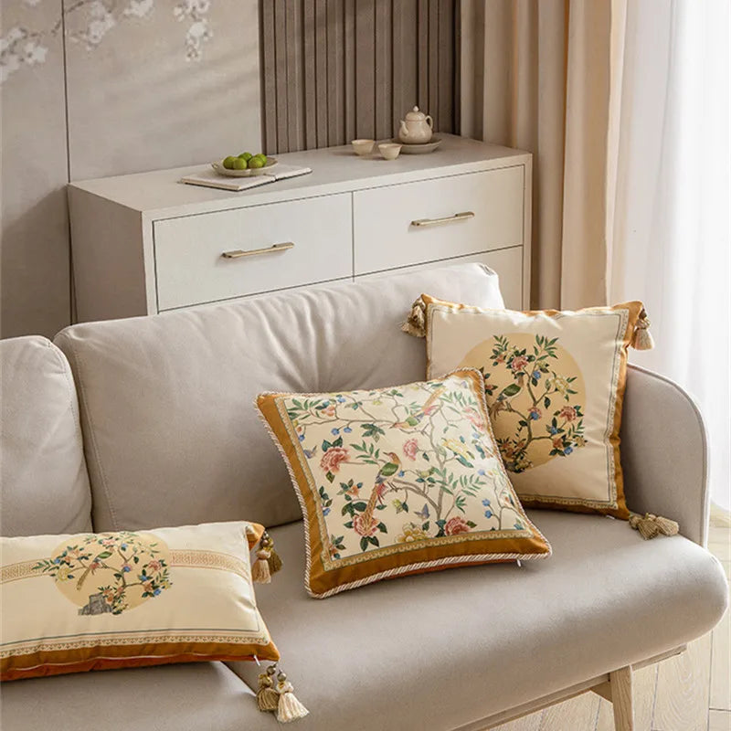 Three "Golden Garden" chinoiserie velvet cushion covers styled on a grey linen sofa in a neutral living room, square and lumbar sizes with exotic birds and peonies, gold braided border and corner tassels — CL House Cushions collection