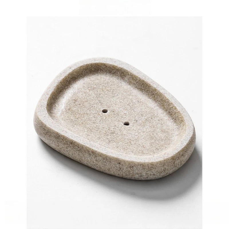 Oval pebble-shaped stone-texture resin soap dish in warm sand beige with two drainage holes, top angle view, white background
