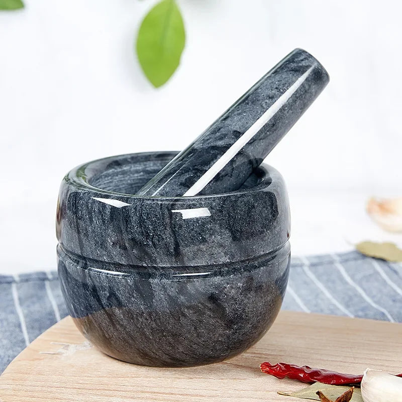 Dark grey marble mortar and pestle set on a wooden chopping board with blue striped linen cloth — garlic cloves, dried red chillies and bay leaves scattered around, green leaf in soft-focus background — lifestyle shot of the Dark Marble variant of the Stone Grind Mortar & Pestle Set by CL House