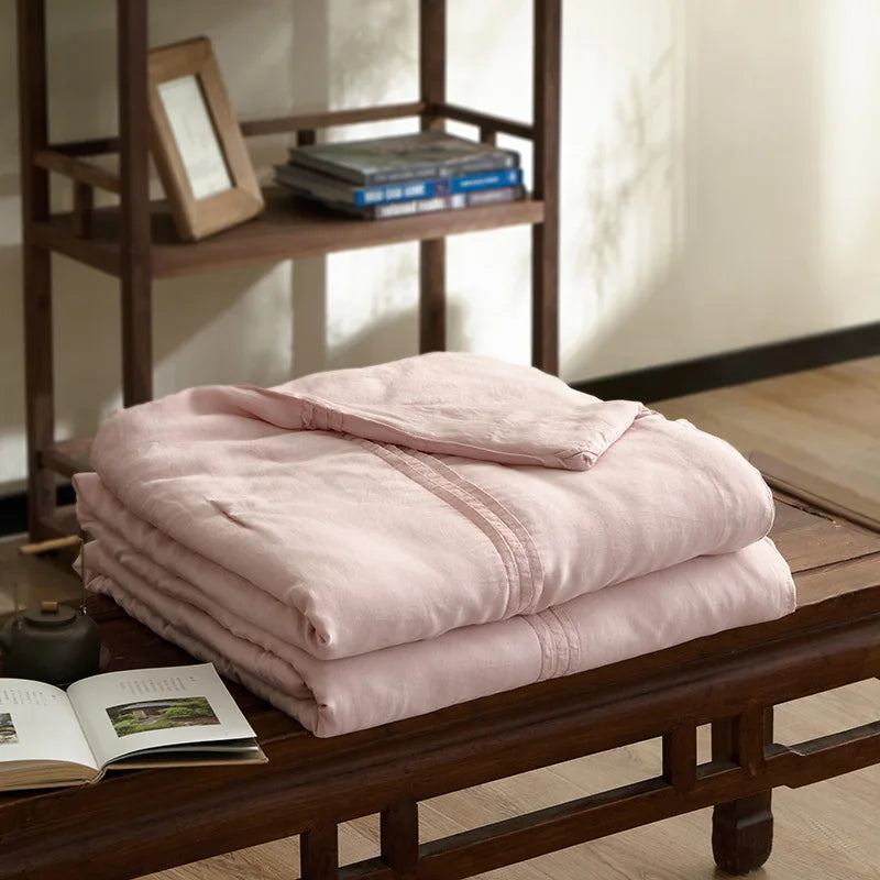 Blush pink quilted linen cotton summer throw blanket, neatly folded on a dark wood bench in a japandi-style interior with wooden shelving and open book