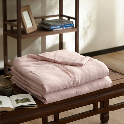 Blush pink quilted linen cotton summer throw blanket, neatly folded on a dark wood bench in a japandi-style interior with wooden shelving and open book