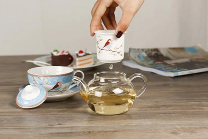 Hand lifting the porcelain infuser cup over a glass teapot filled with golden herbal tea, with a blue "Happy Bird" cup and saucer on a wooden surface, cake and magazine in the background.