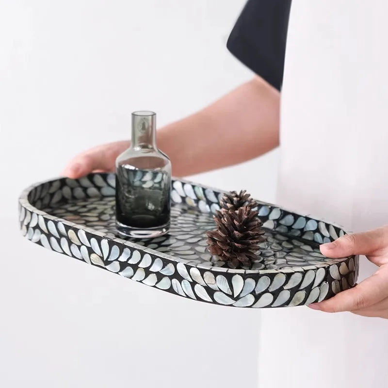  Hands holding midnight black mother-of-pearl oval tray styled with smoky glass bottle and pine cone, iridescent blue-grey shell mosaic detail — artisan bathroom tray lifestyle