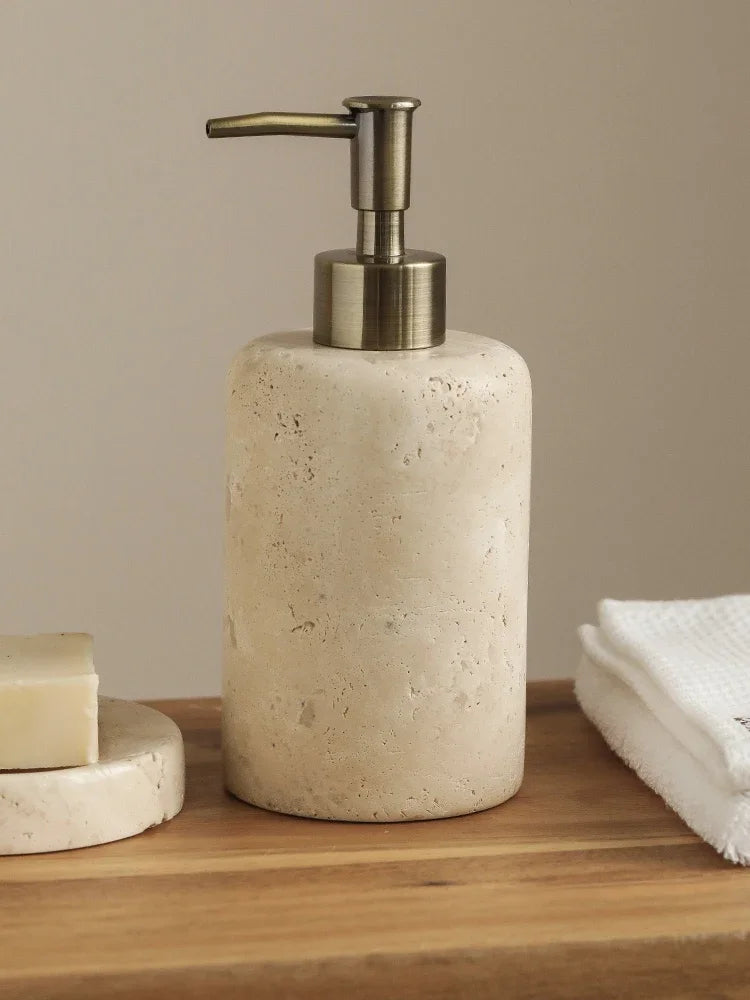 Natural travertine soap dispenser with antique brass pump on a wooden shelf, warm beige porous stone texture, soap dish with bar soap alongside — wabi-sabi bathroom detail