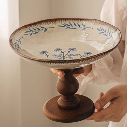  "Blue Botanics" Blue Freesia ceramic pedestal serving stand held by two hands, showing scale and the hand-painted botanical motif with solid walnut wood base — lifestyle shot