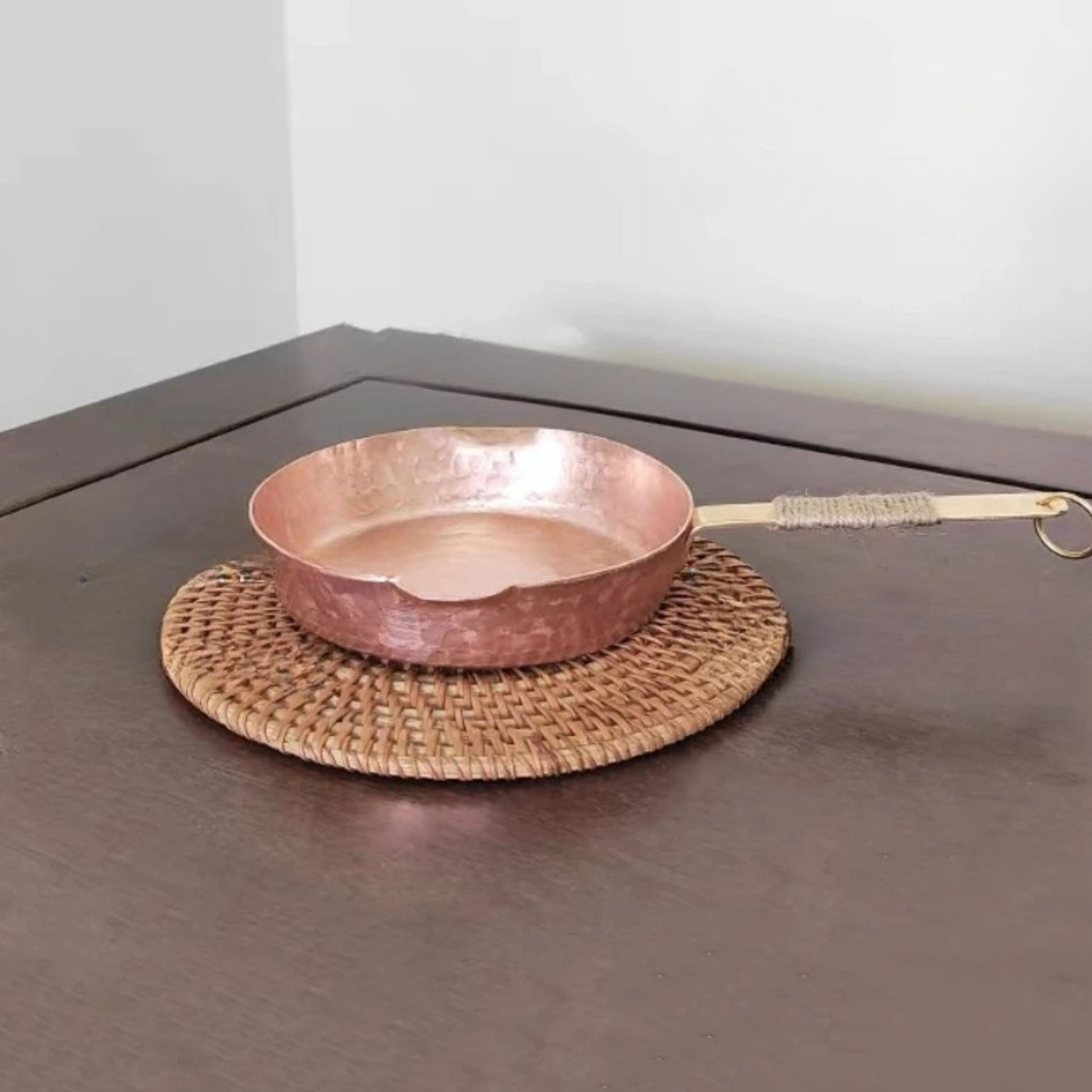 Copper Skillet handmade pure copper frying pan on a round rattan trivet placed on a dark wood surface, showing the hammered rose-gold body and brass jute-wrapped handle with ring hook