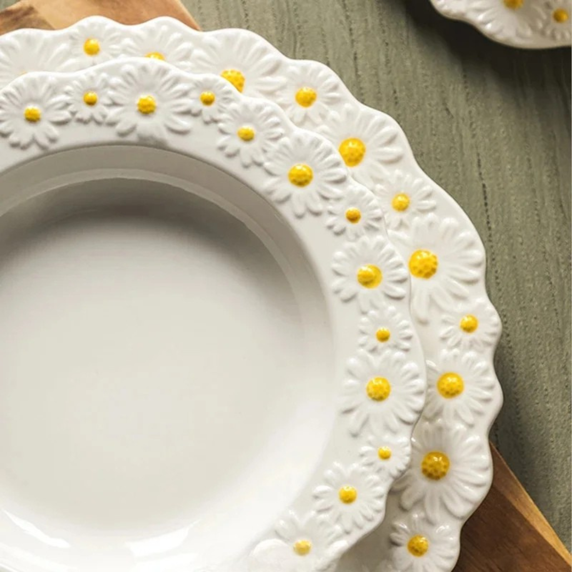 "Daisy Bloom" ceramic dinner plate — close-up detail of raised daisy relief border with white petals and golden yellow centres on grey wood surface, showing depth and texture of the scalloped edge — lifestyle detail shot