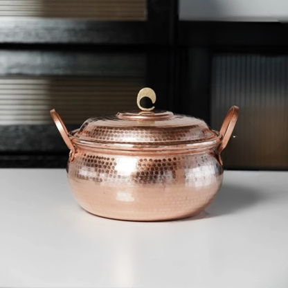 Copper Cauldron handmade pure copper stew pot with lid styled on a white kitchen counter with dark glass cabinet doors in the background, showing the full rose-gold hammer-textured body and brass crescent lid knob