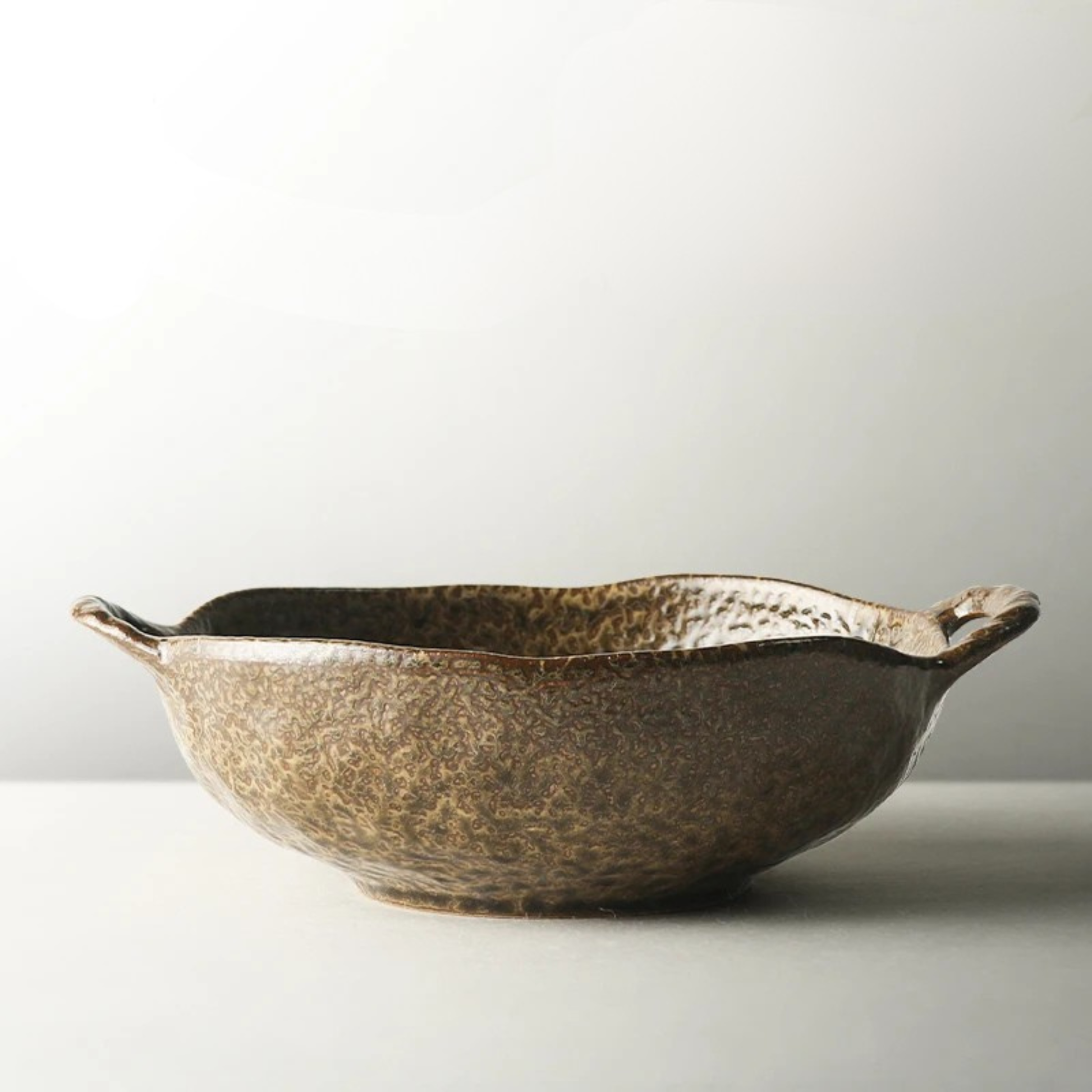 "Wabi Crest" stoneware serving bowl in warm earth brown with speckled reactive glaze and organic rippled rim — side view on white background