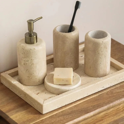  Natural travertine bathroom set styled on a wooden surface — soap dispenser with brass pump, toothbrush cup, mouthwash cup, round soap dish and rectangular tray in warm beige stone — japandi bathroom decor