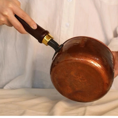  Hand tilting the Copper Forge saucepan to show the hammered pure copper base — dark wooden handle with brass fitting visible, warm copper patina covering the entire exterior including the flat bottom, white linen cloth background — craftsmanship detail shot by CL House