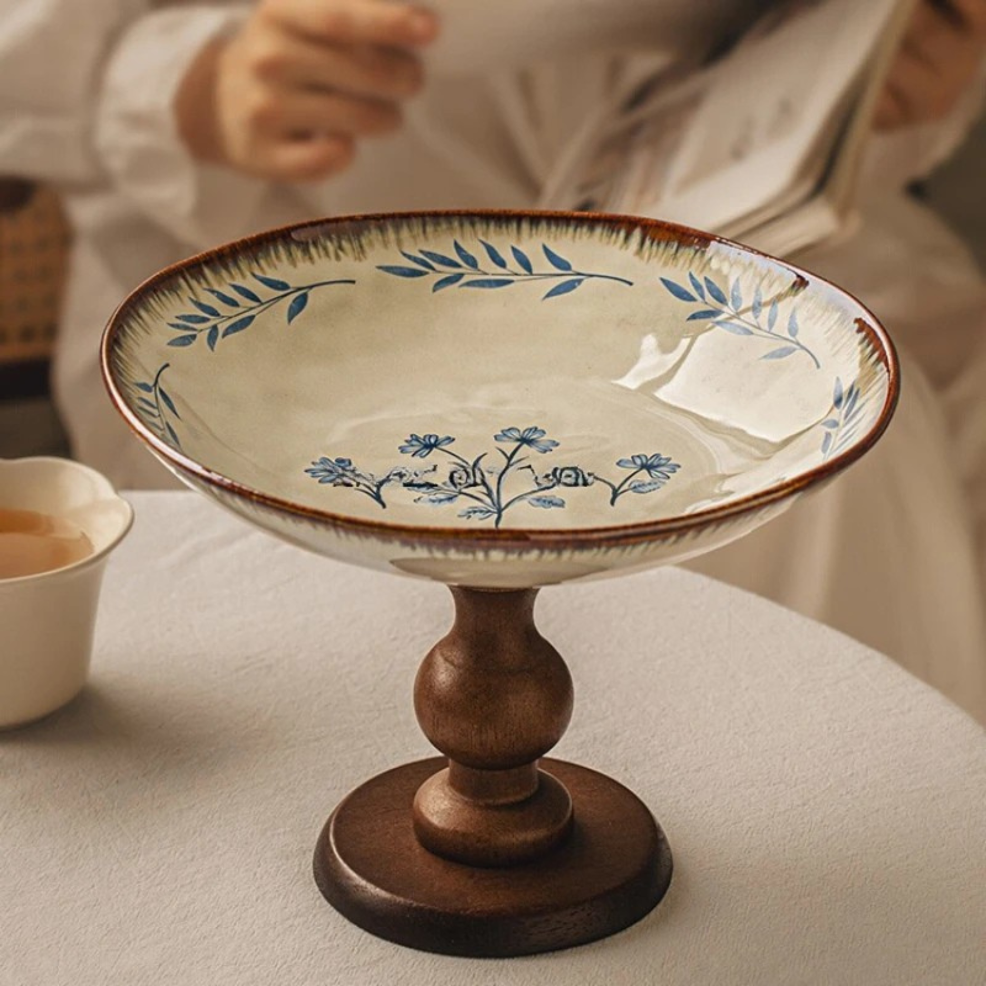"Blue Botanics" Blue Freesia ceramic pedestal serving stand on a linen-covered table, styled with a tea cup — lifestyle shot with soft natural light