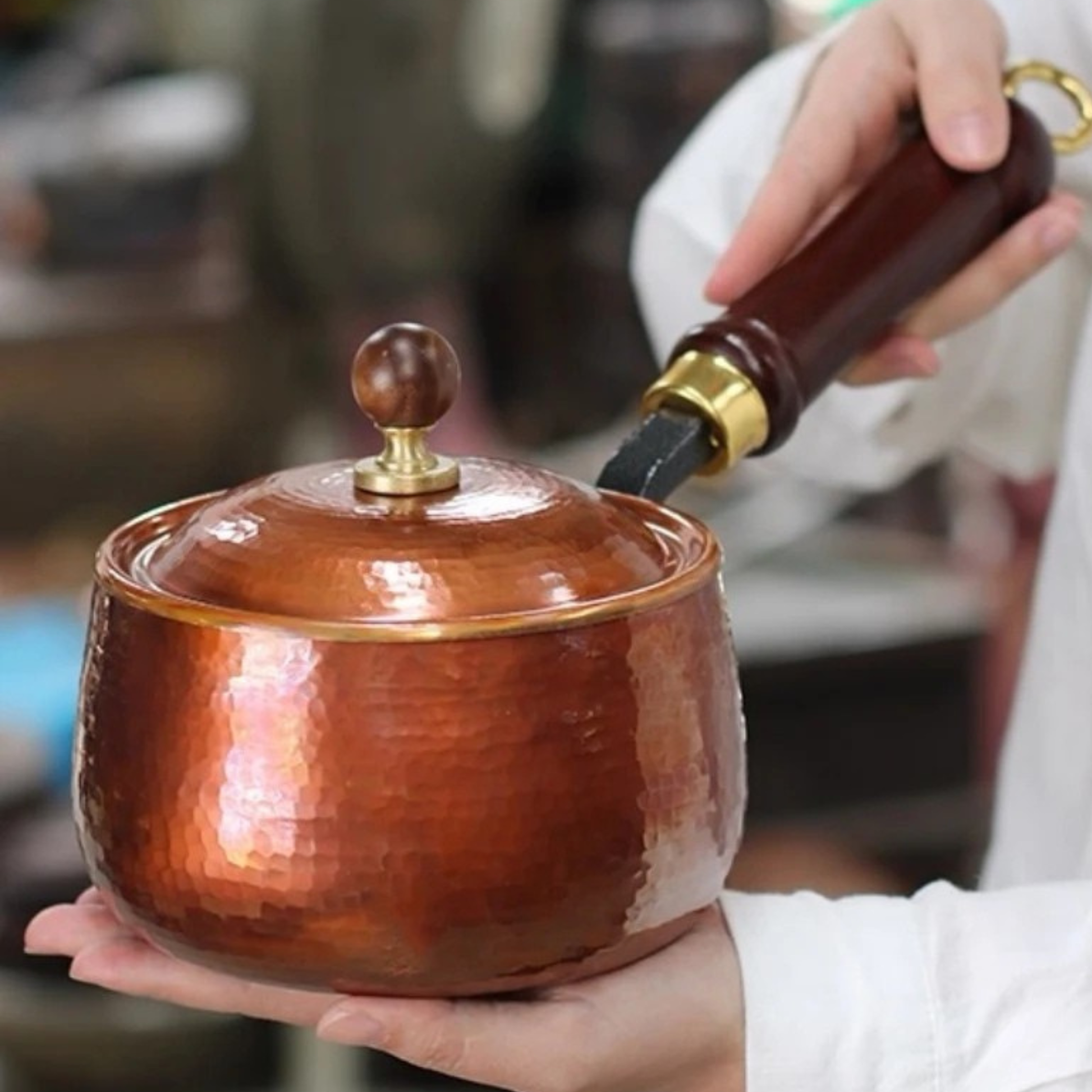  Two hands holding the closed Copper Forge saucepan in a professional kitchen setting — hammered copper body with brass rim, domed lid with wooden knob and dark wooden handle with brass fitting clearly visible, blurred stainless steel kitchen background — scale and craftsmanship lifestyle shot by CL House