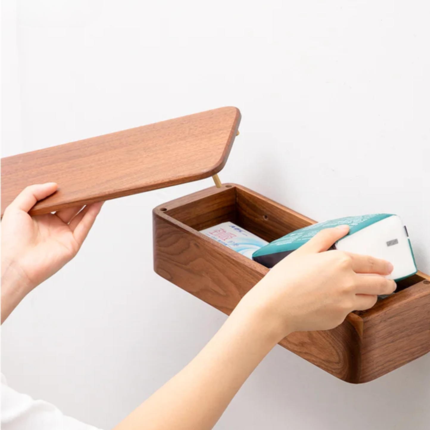  Hands opening the hinged lid of a walnut wood wall-mounted tissue holder revealing interior storage compartment with tissue packets — functional detail view