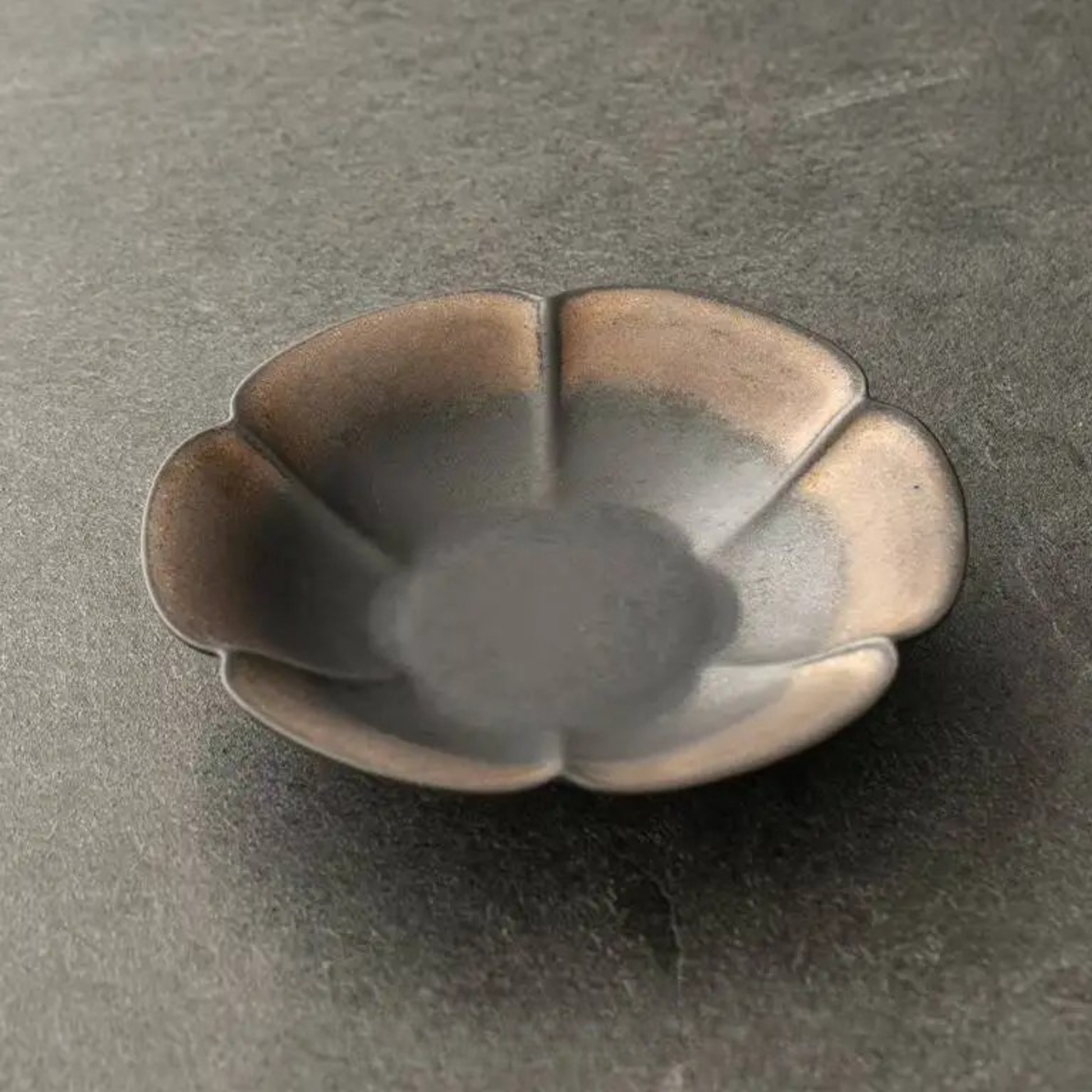 Charcoal petal ceramic dish close-up showing dark matte glaze with bronze rim highlights, dark background