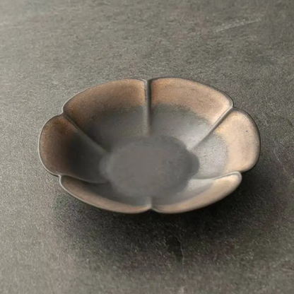 Charcoal petal ceramic dish close-up showing dark matte glaze with bronze rim highlights, dark background