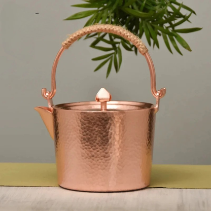 Copper Ritual handmade pure copper teapot kettle in Rose Gold finish styled on a linen table runner with a bamboo plant in the background, showing the full hammer-textured body and jute-wrapped arched handle